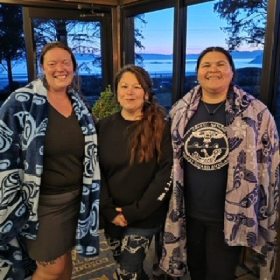 Three people standing indoors, wrapped in blankets, with a view of the beach through windows.