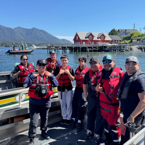 Group of people in life vests on a boat with a harbor and houses in the background.