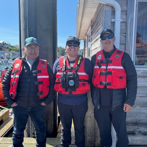 Three people in red life vests smiling on a dock near a wooden building.