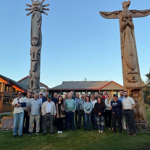 Group of people posing between two large wooden totem poles, with a building in the background.