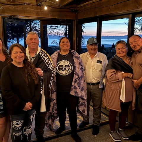 Group of seven people smiling indoors with a scenic coastal view in the background.