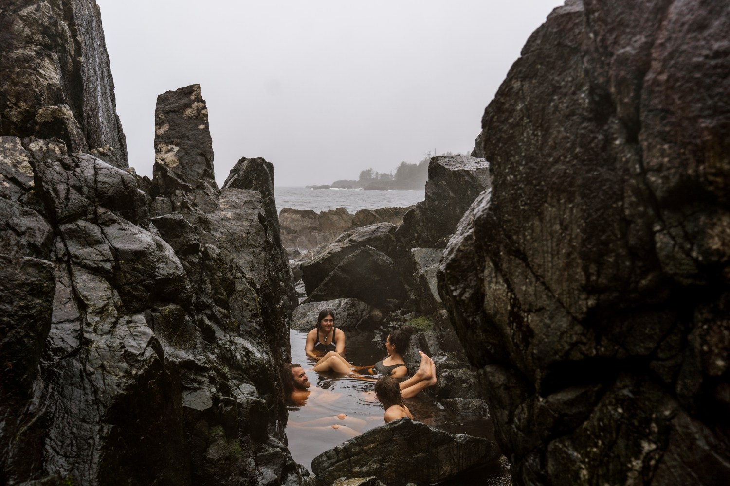 Four people relax in a natural rocky tide pool by the sea on a cloudy day.