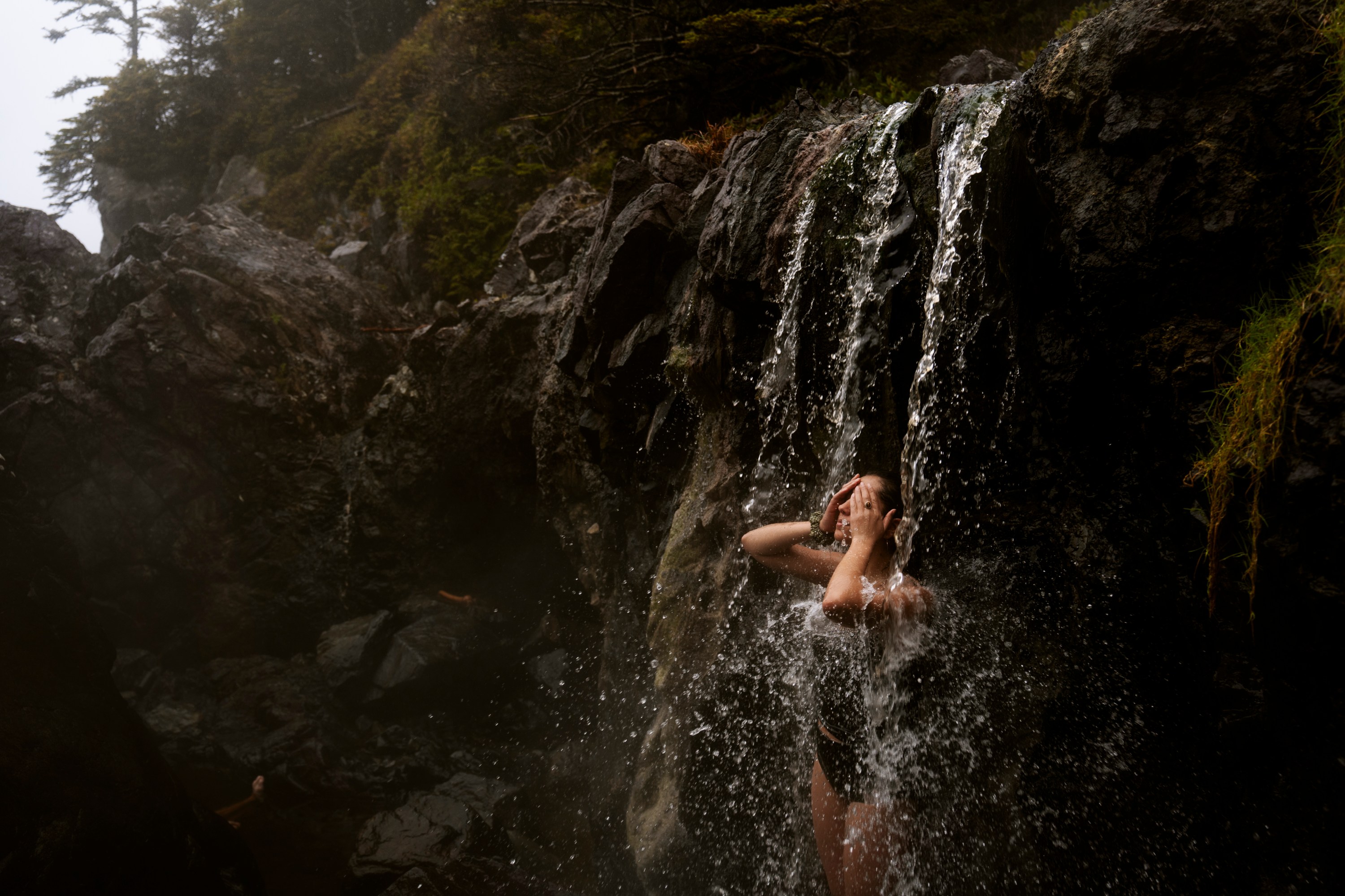 Person showering under a small waterfall surrounded by rocks and vegetation.